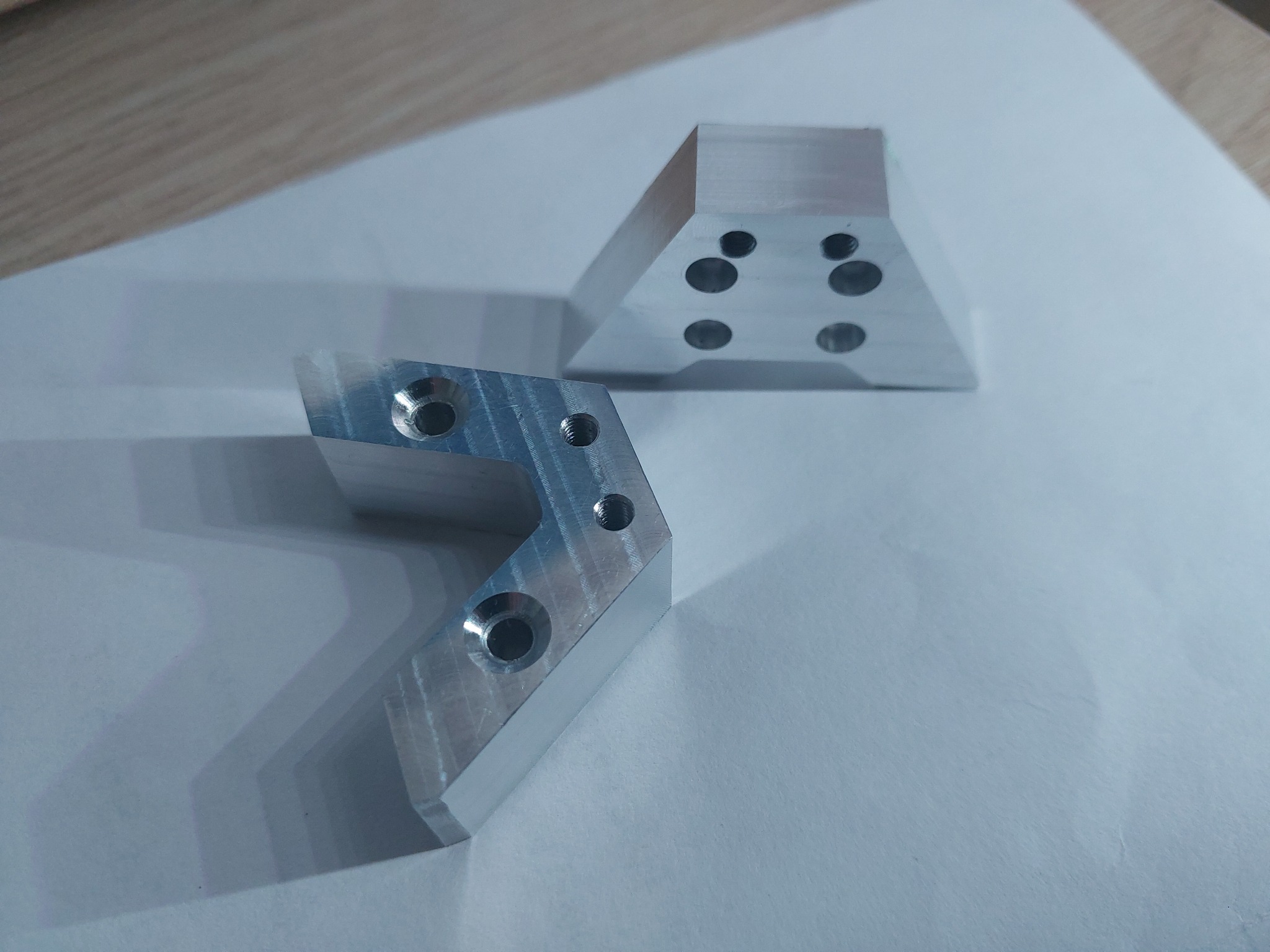 Dwa precyzyjnie wykonane, aluminiowe elementy z otworami, leżące na białym tle. Jeden w kształcie litery L, drugi trapezoidalny. Widoczne ślady obróbki.