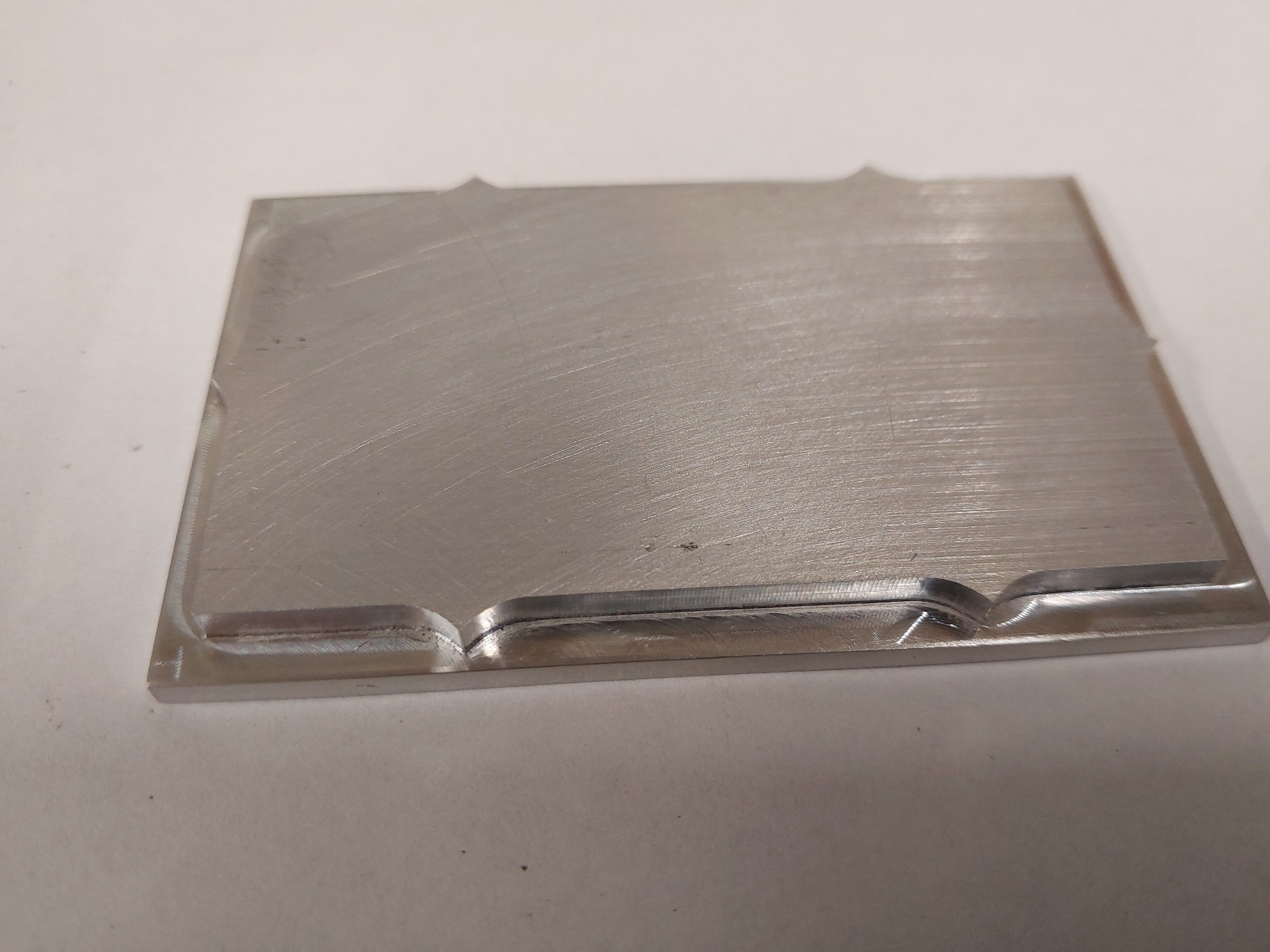 Szlifowany, prostokątny element metalowy z precyzyjnie wyfrezowanymi wgłębieniami na krawędziach, leżący na jasnym tle. Widoczne ślady obróbki.