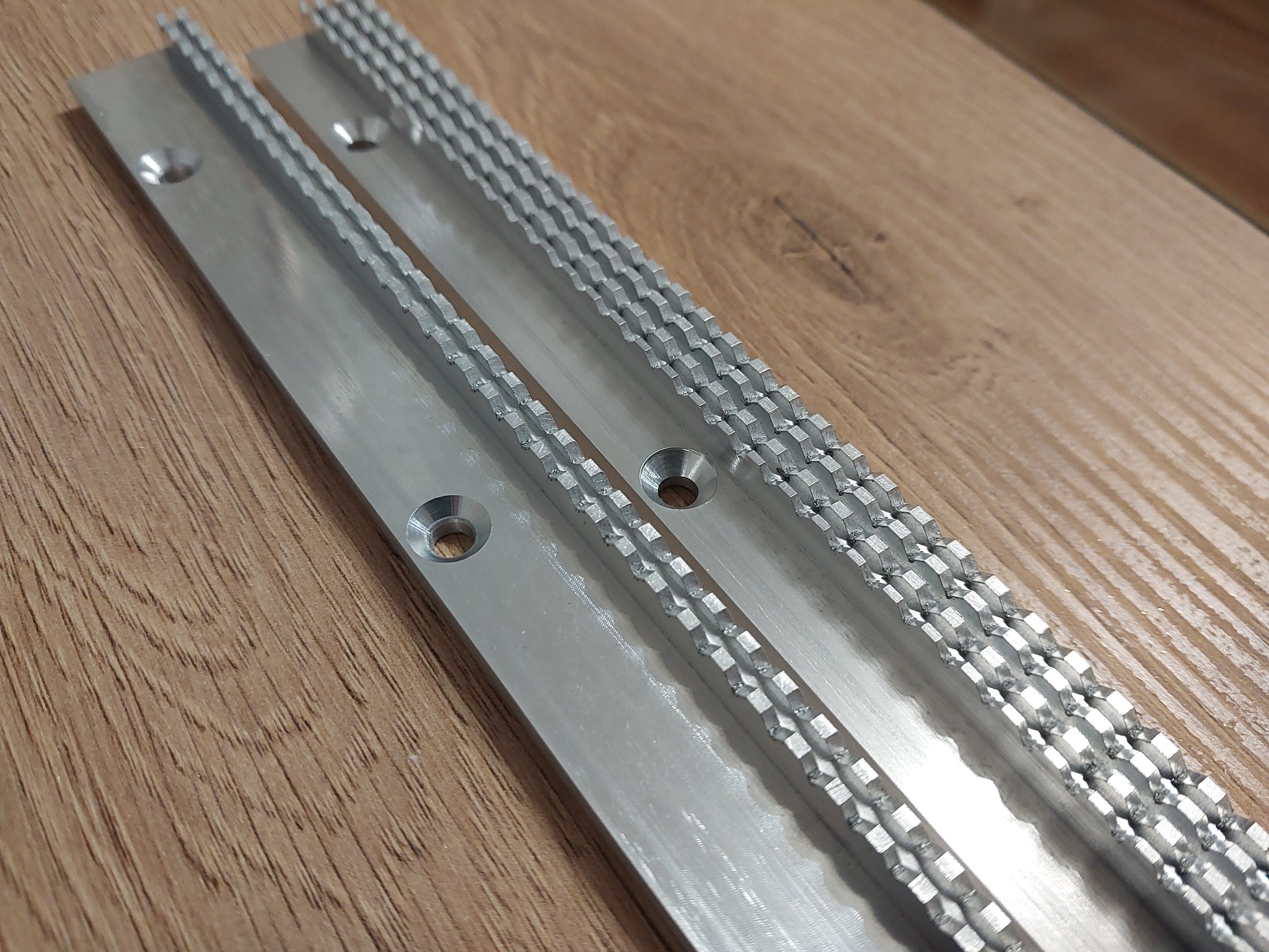 Dwie aluminiowe listwy z precyzyjnie frezowanymi zębami, leżące ukośnie na drewnianej powierzchni. Widoczne otwory montażowe i detale obróbki.