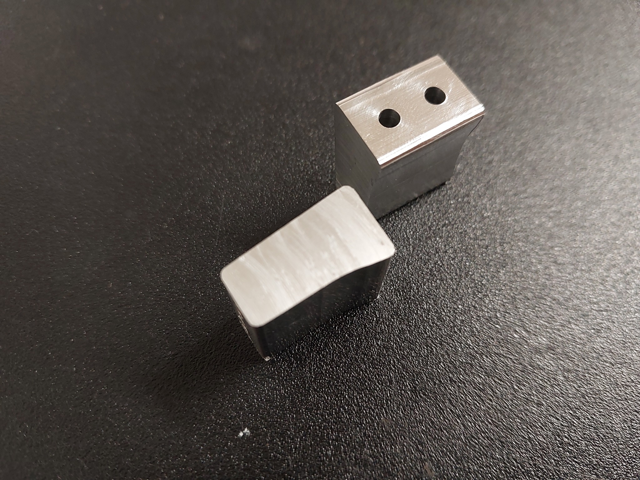 Dwa precyzyjnie obrobione, metalowe prostopadłościany z otworami na ciemnym tle. Jeden leży, drugi stoi, ukazując detale obróbki CNC.