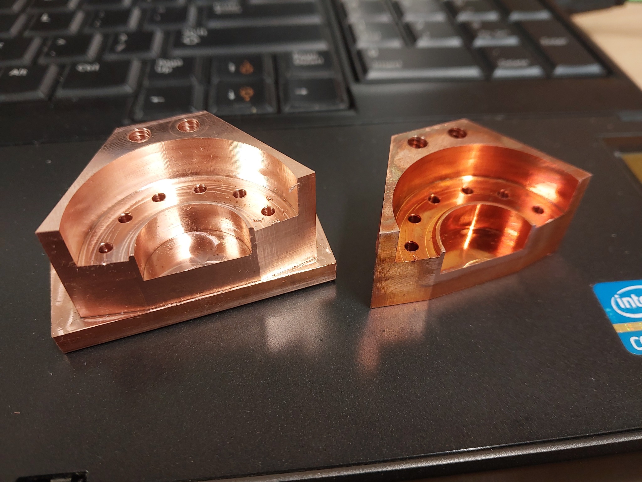 Dwie precyzyjnie obrobione, miedziane części z otworami, leżące na laptopie. Widoczne ślady obróbki skrawaniem i detale wykonania.