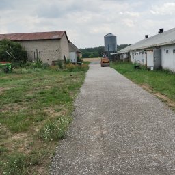 Krzyś-Kop - Walec drogowy ubija żwirową drogę między budynkami gospodarczymi. W tle silos i pochmurne niebo. Widok na utwardzoną drogę z perspektywy pieszego.