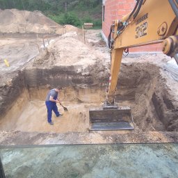 Krzyś-Kop - Żółta koparka Liebherr 312 w wykopie, operator wyrównuje dno łopatą. Widok z kabiny, w tle ceglany budynek w budowie i hałda ziemi.