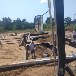 Krzyś-Kop - Fundamenty domu w trakcie budowy. Pracownicy zagęszczają piasek ubijarkami, w tle koparka. Widok z kabiny maszyny budowlanej, z lustrem wstecznym.