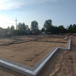 Krzyś-Kop - Fundamenty domu jednorodzinnego w trakcie budowy, widoczna wykopana ziemia, szalunki i zbrojenia. W tle koparka i częściowo widoczny budynek.