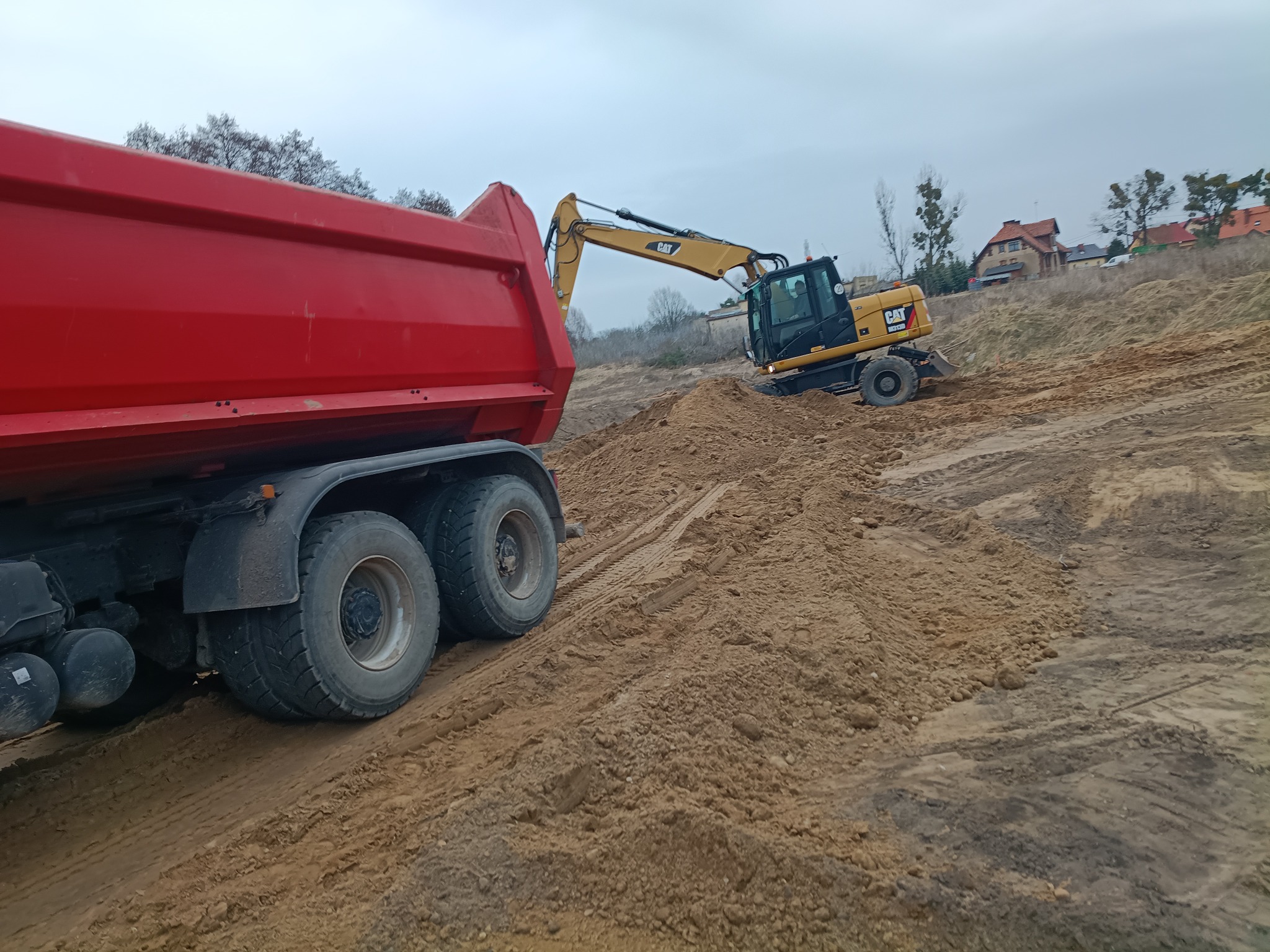 Czerwona wywrotka i żółta koparka CAT na placu budowy. Koparka wyrównuje teren, w tle zabudowa jednorodzinna i pochmurne niebo. Widoczne ślady opon na ziemi.