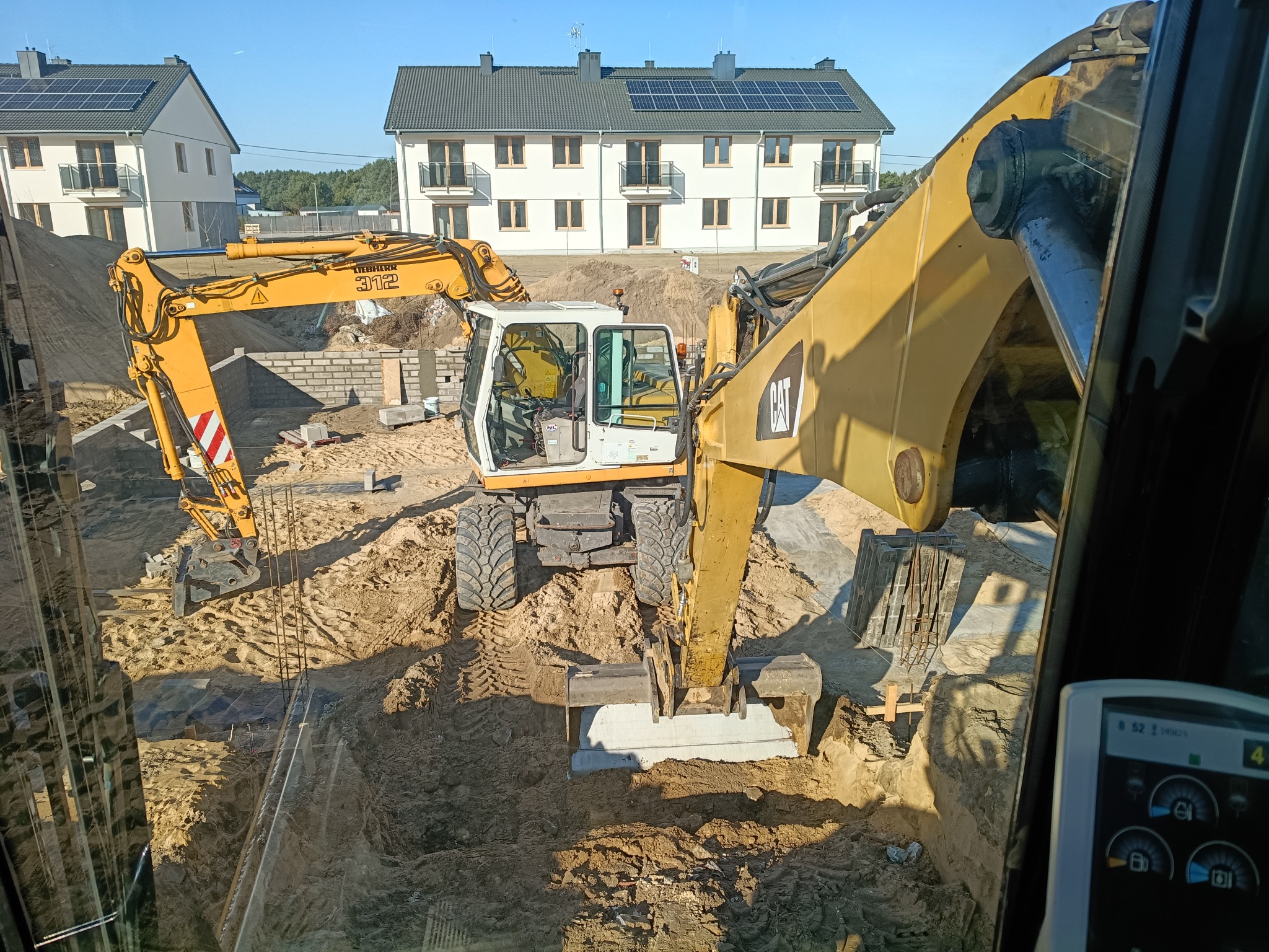 Żółta koparka CAT i Liebherr na placu budowy z widokiem na nowe domy z panelami słonecznymi, widziana z kabiny operatora. Wykop pod fundamenty.