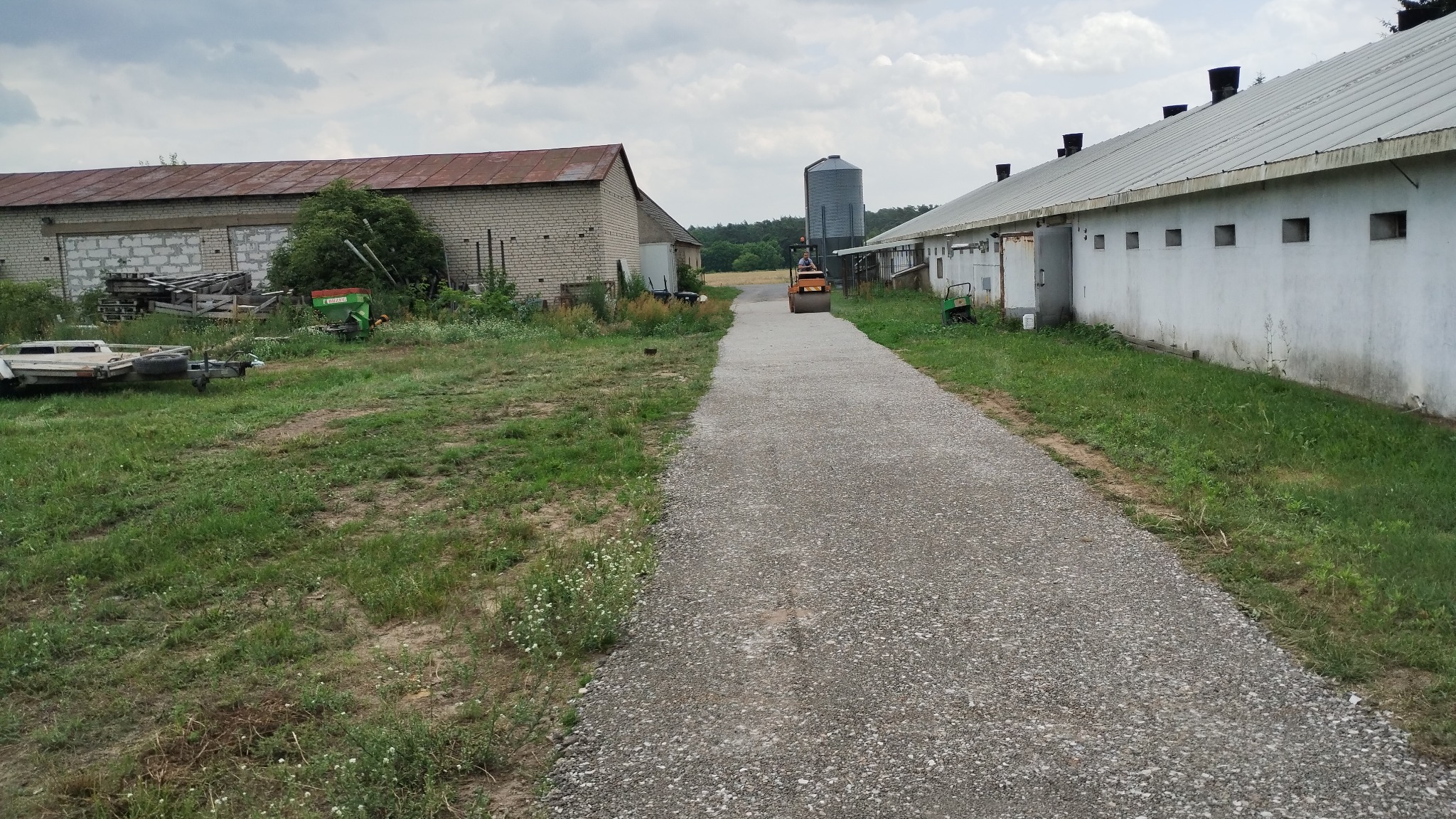Walec drogowy ubija żwirową drogę między budynkami gospodarczymi. W tle silos i pochmurne niebo. Widok na utwardzoną drogę z perspektywy pieszego.