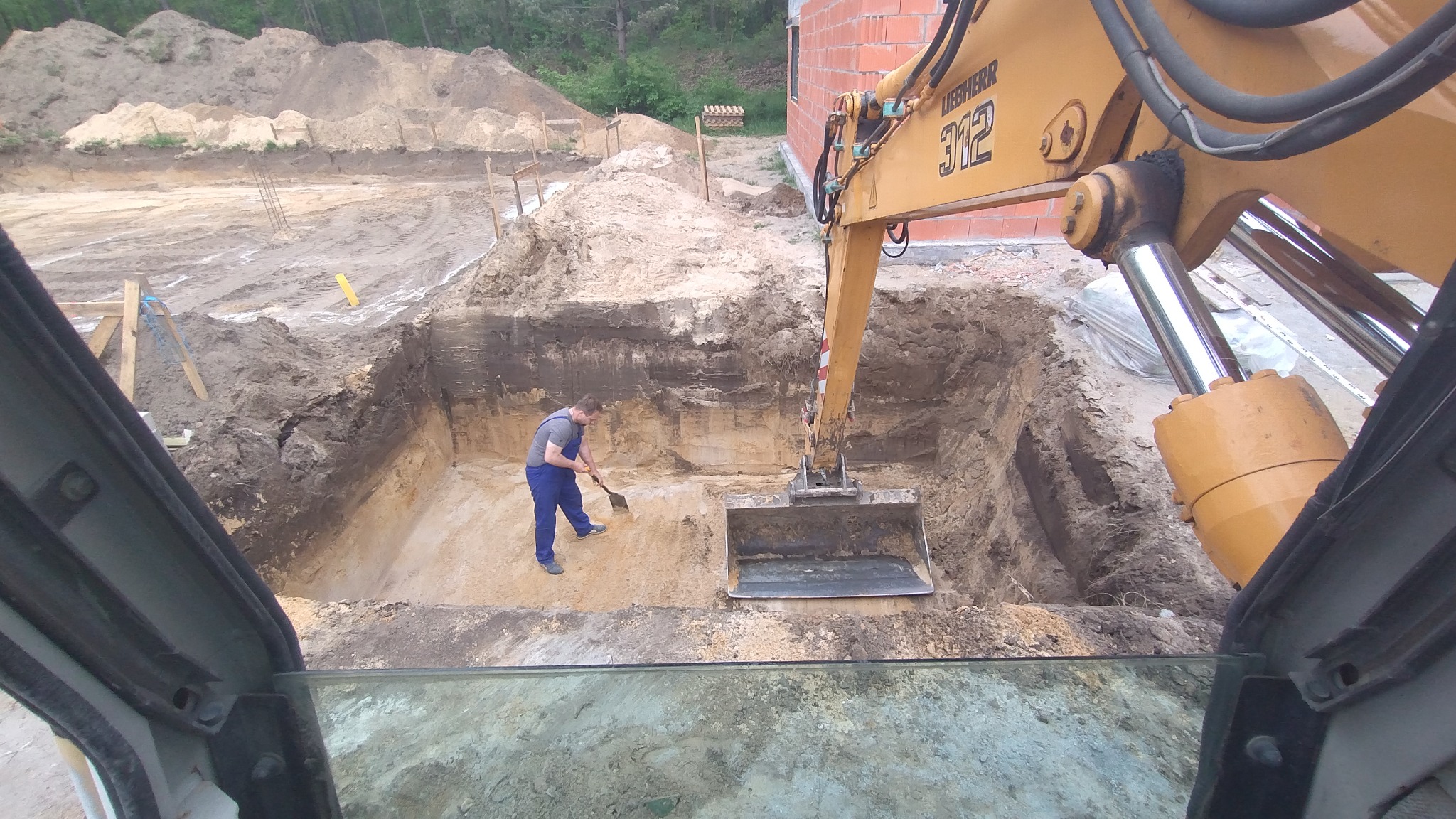 Żółta koparka Liebherr 312 w wykopie, operator wyrównuje dno łopatą. Widok z kabiny, w tle ceglany budynek w budowie i hałda ziemi.
