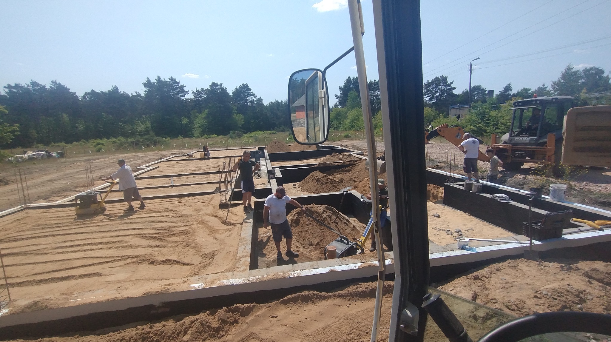 Fundamenty domu w trakcie budowy. Pracownicy zagęszczają piasek ubijarkami, w tle koparka. Widok z kabiny maszyny budowlanej, z lustrem wstecznym.