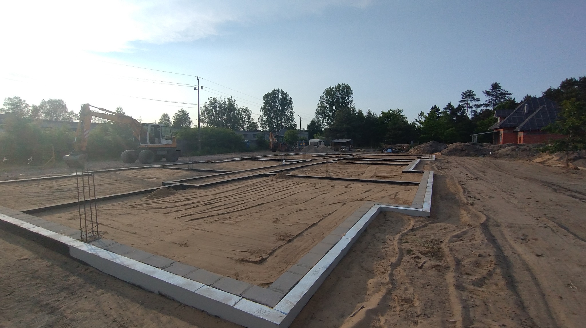 Fundamenty domu jednorodzinnego w trakcie budowy, widoczna wykopana ziemia, szalunki i zbrojenia. W tle koparka i częściowo widoczny budynek.
