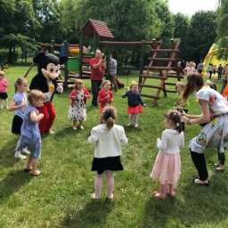 Animacje dla dzieci - Animator w stroju Myszki Miki bawi grupę dzieci na zielonej trawie, w tle plac zabaw i dorośli. Ujęcie z perspektywy dziecka, z bliska, w słoneczny dzień.