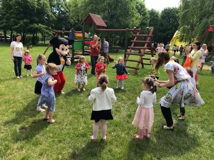 Animator w stroju Myszki Miki bawi grupę dzieci na zielonej trawie, w tle plac zabaw i dorośli. Ujęcie z perspektywy dziecka, z bliska, w słoneczny dzień.