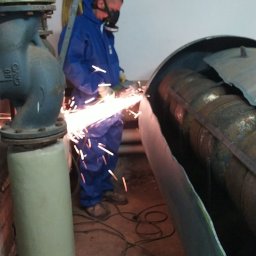 Profesjonalne usługi rozbiórek ręcznych Michał K. - Pracownik w kombinezonie i masce tnie metalową rurę szlifierką kątową, iskry. W tle instalacja przemysłowa i fragment ściany. Praca w pomieszczeniu.