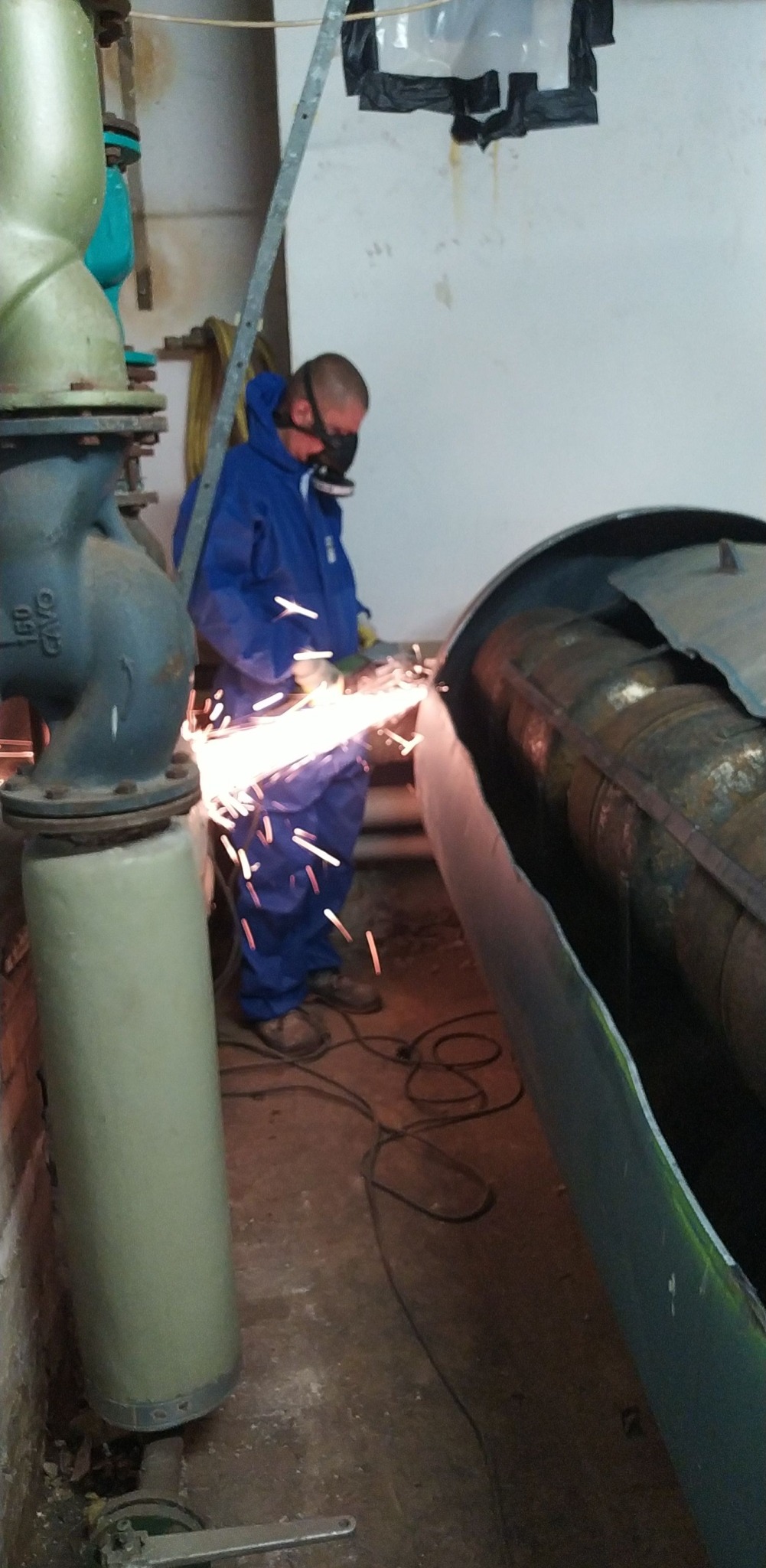 Pracownik w kombinezonie i masce tnie metalową rurę szlifierką kątową, iskry. W tle instalacja przemysłowa i fragment ściany. Praca w pomieszczeniu.