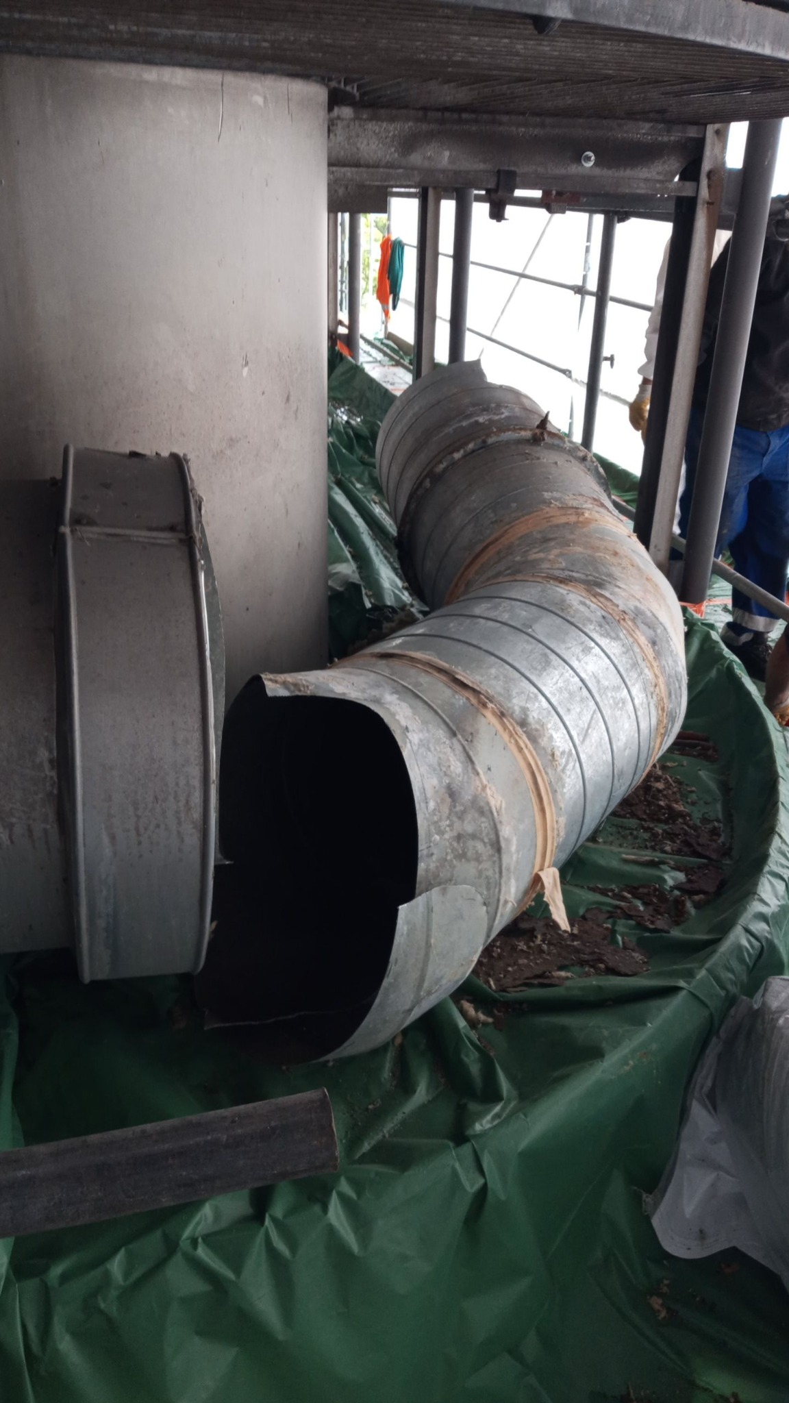 Fragmenty metalowych rur wentylacyjnych na zielonej folii budowlanej, pozostałości po demontażu instalacji wewnątrz budynku. Widoczne elementy konstrukcji stropu.