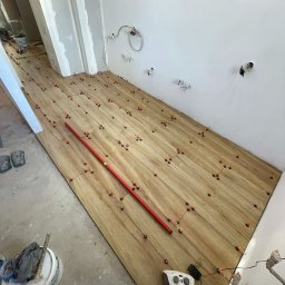 Usługi Budowlane Mikołaj Rosoliński - Układanie drewnopodobnych płytek na podłodze z użyciem poziomicy i systemu poziomowania. Widoczne narzędzia i akcesoria glazurnicze. Ściany przygotowane do dalszych prac.