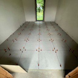 Usługi Budowlane Mikołaj Rosoliński - Szare płytki podłogowe w trakcie układania, z pomarańczowymi krzyżykami dystansowymi, widok z góry na surowy stan pomieszczenia z oknem.
