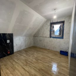 Usługi Budowlane Mikołaj Rosoliński - Wykończona łazienka na poddaszu z drewnopodobną podłogą, marmurowymi płytkami ściennymi i czarną zabudową. Okno zabezpieczone folią. Widoczne narzędzia glazurnicze.