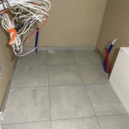 Usługi Budowlane Mikołaj Rosoliński - Ułożone szare płytki podłogowe w małym pomieszczeniu z wystającymi przewodami i rurami instalacyjnymi. Widok z góry, surowy stan przed wykończeniem.