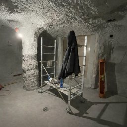 Usługi Budowlane Mikołaj Rosoliński - Surowe wnętrze z fakturą na ścianach i suficie. Metalowe rusztowanie z kurtką w centrum. Widoczne elementy instalacji elektrycznej i otwór w suficie.