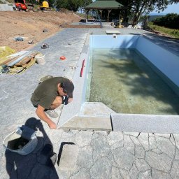 Usługi Budowlane Mikołaj Rosoliński - Pracownik w trakcie układania kamiennej mozaiki wokół basenu zewnętrznego. W tle widoczny krajobraz z drzewami i budynkami. Na pierwszym planie narzędzia pracy.