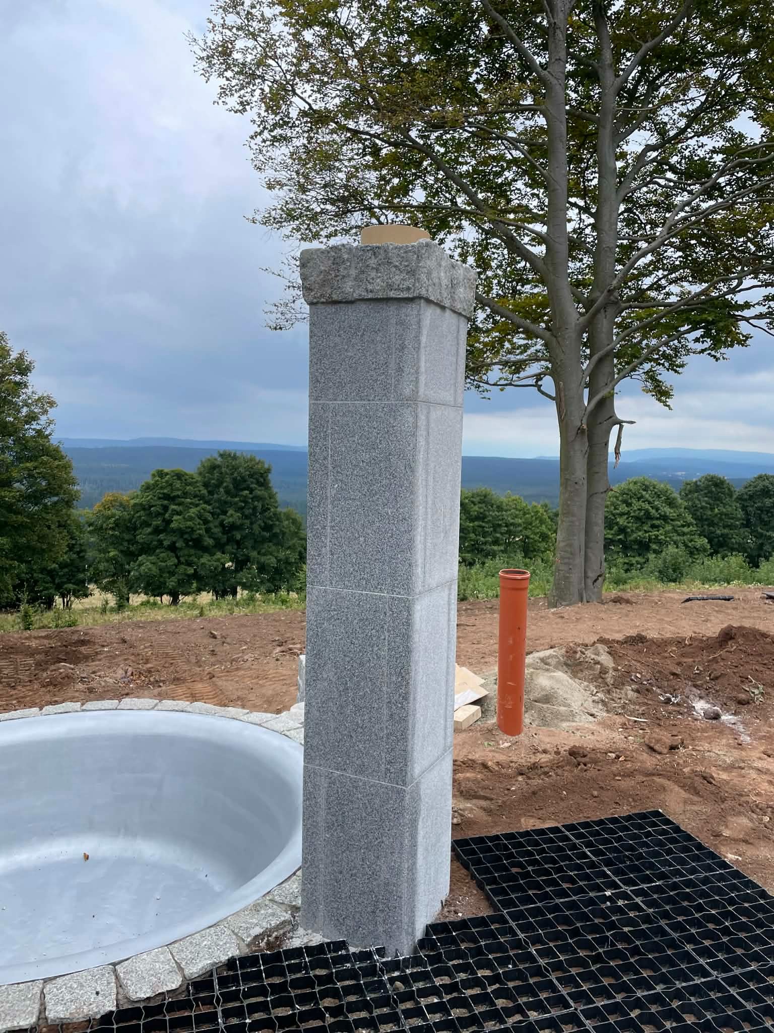 Granitowa kolumna z obudową basenu w tle krajobrazu. Widoczne elementy instalacji i przygotowania terenu. Ujęcie z bliska, podkreślające detale kamieniarskie.