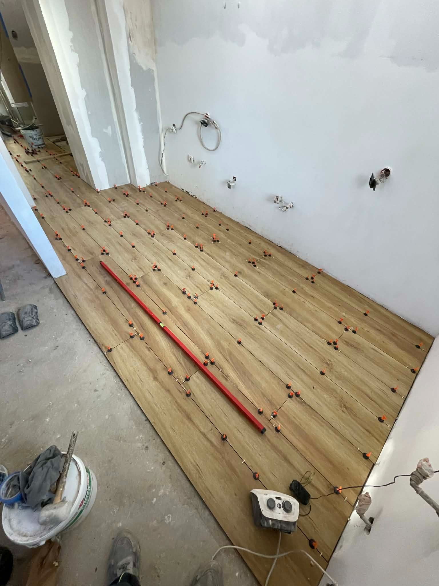 Układanie drewnopodobnych płytek na podłodze z użyciem poziomicy i systemu poziomowania. Widoczne narzędzia i akcesoria glazurnicze. Ściany przygotowane do dalszych prac.