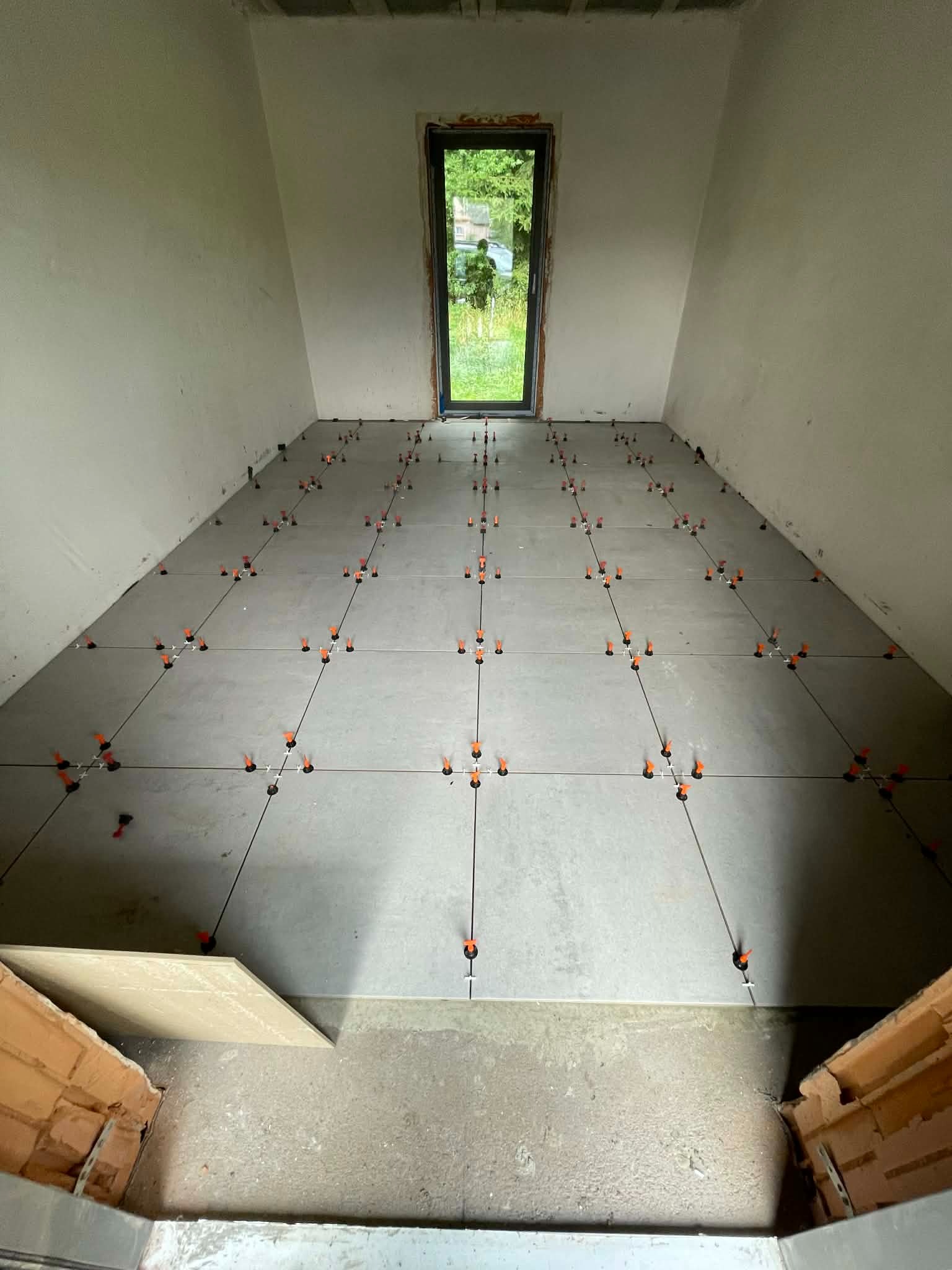 Szare płytki podłogowe w trakcie układania, z pomarańczowymi krzyżykami dystansowymi, widok z góry na surowy stan pomieszczenia z oknem.