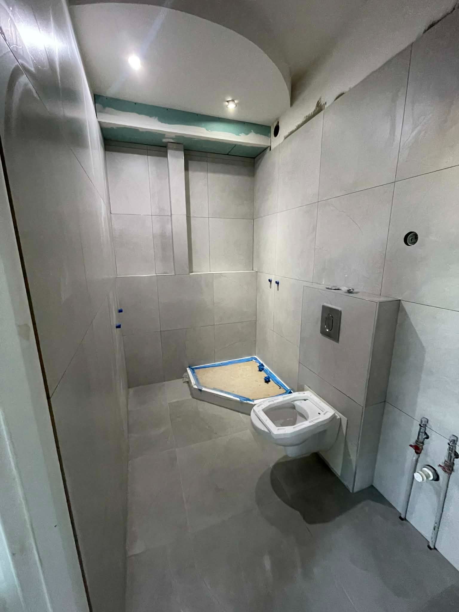 Szara łazienka w trakcie remontu: ułożone płytki, zamontowana toaleta, widoczne elementy instalacji. Fragment sufitu podwieszanego z płyt gipsowych.