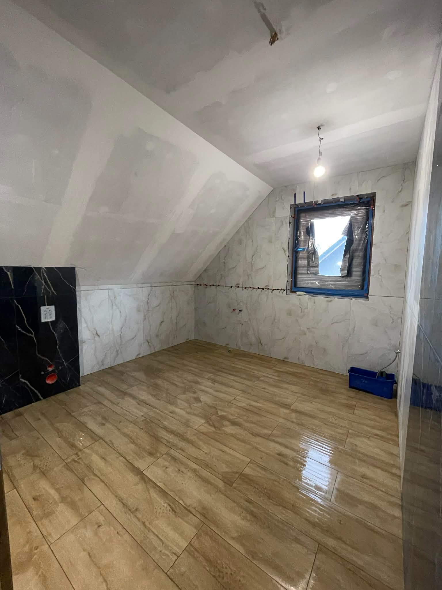 Wykończona łazienka na poddaszu z drewnopodobną podłogą, marmurowymi płytkami ściennymi i czarną zabudową. Okno zabezpieczone folią. Widoczne narzędzia glazurnicze.