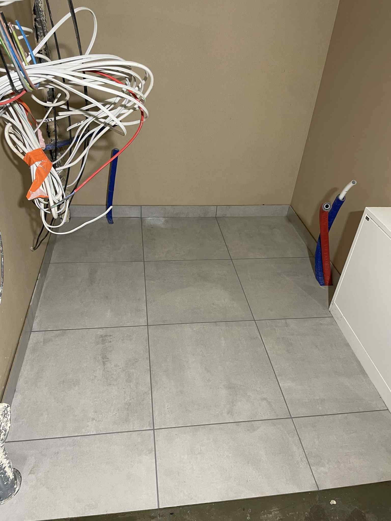 Ułożone szare płytki podłogowe w małym pomieszczeniu z wystającymi przewodami i rurami instalacyjnymi. Widok z góry, surowy stan przed wykończeniem.