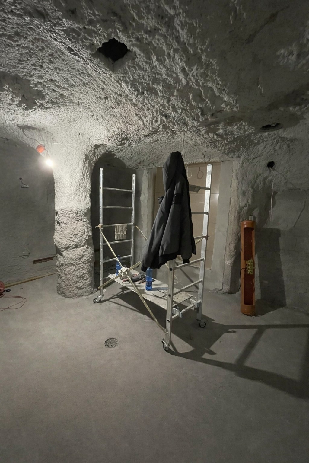 Surowe wnętrze z fakturą na ścianach i suficie. Metalowe rusztowanie z kurtką w centrum. Widoczne elementy instalacji elektrycznej i otwór w suficie.