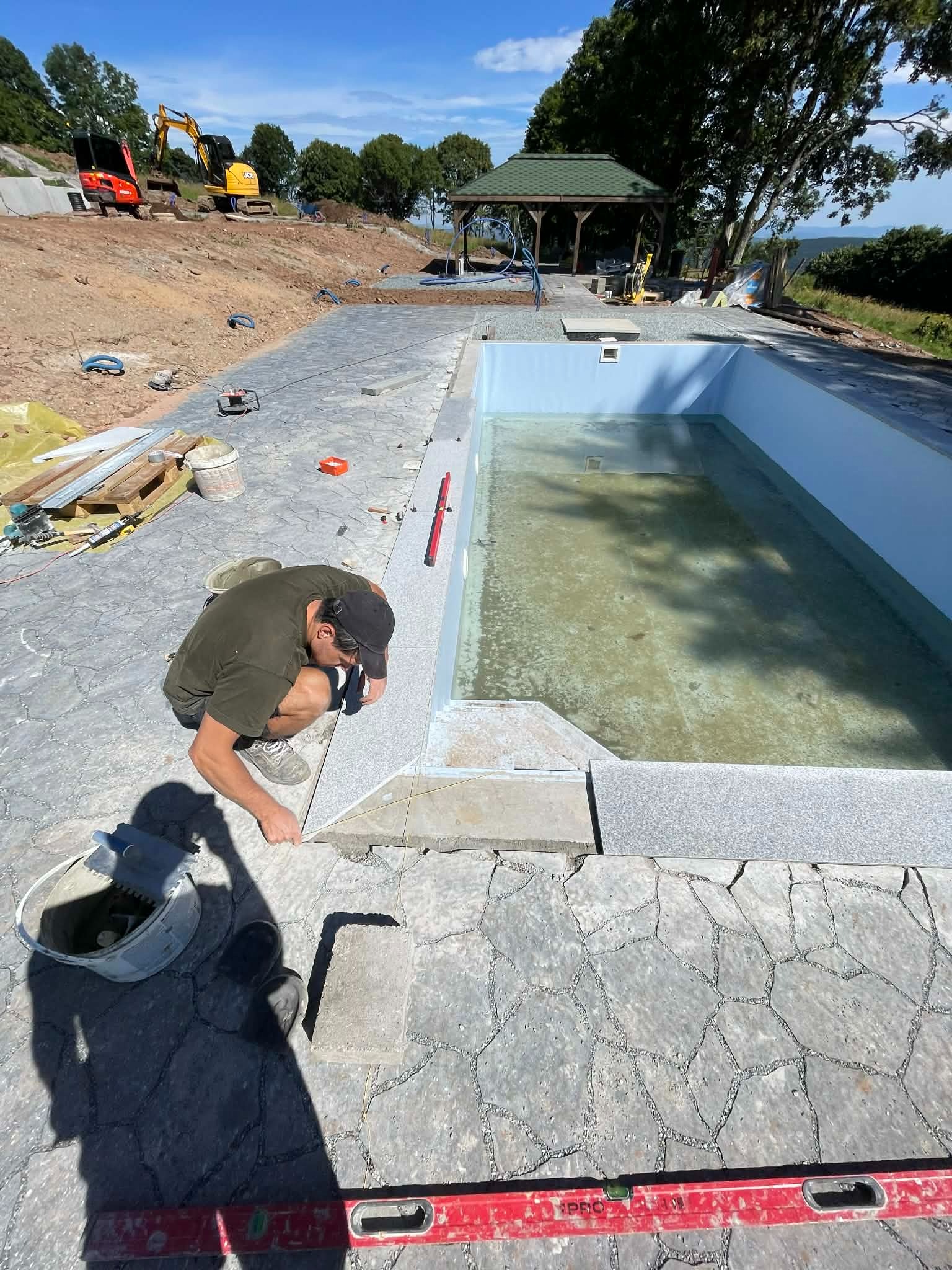 Pracownik w trakcie układania kamiennej mozaiki wokół basenu zewnętrznego. W tle widoczny krajobraz z drzewami i budynkami. Na pierwszym planie narzędzia pracy.