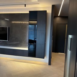 InStyle - Nowoczesny salon z parkietem w jodełkę, ciemną zabudową ściany z podświetleniem LED i widokiem na kolejne pomieszczenie przez otwarte drzwi.