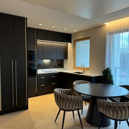 InStyle - Nowoczesna kuchnia z ciemnymi szafkami, złotymi akcentami i parkietem w jodełkę. Okrągły stół z krzesłami w stylu glamour. Minimalistyczne oświetlenie.