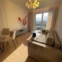 InStyle - Jasne wnętrze salonu z beżową sofą, stolikiem kawowym i krzesłami. Widok na miasto przez okno balkonowe. Dekoracyjne listwy na ścianie i nowoczesna lampa.