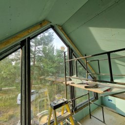 InStyle - Montaż zielonej płyty karton-gips na poddaszu z oknem, widoczne rusztowanie i drabina. Prace wykończeniowe w nowym domu z widokiem na las.