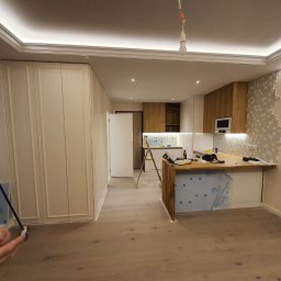 InStyle - Wnętrze w trakcie prac: częściowo ułożone heksagonalne płytki na ścianie, zabudowa kuchenna, drewniany blat, zabezpieczona folią. Widoczny fragment pomalowanej ściany.