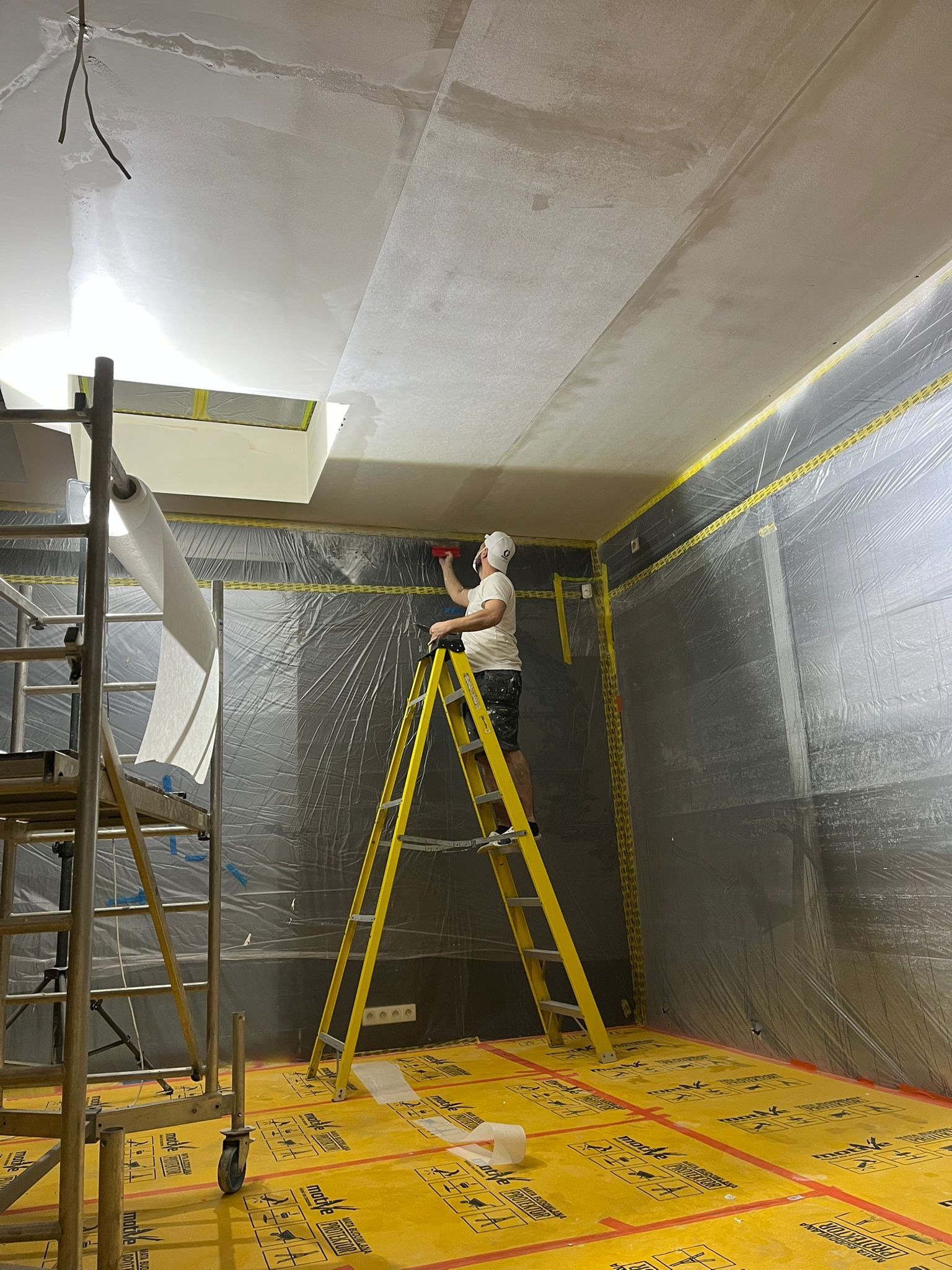 Pracownik na drabinie, w kasku, maluje sufit w pomieszczeniu zabezpieczonym folią i żółtą matą ochronną. Widoczny fragment rusztowania.