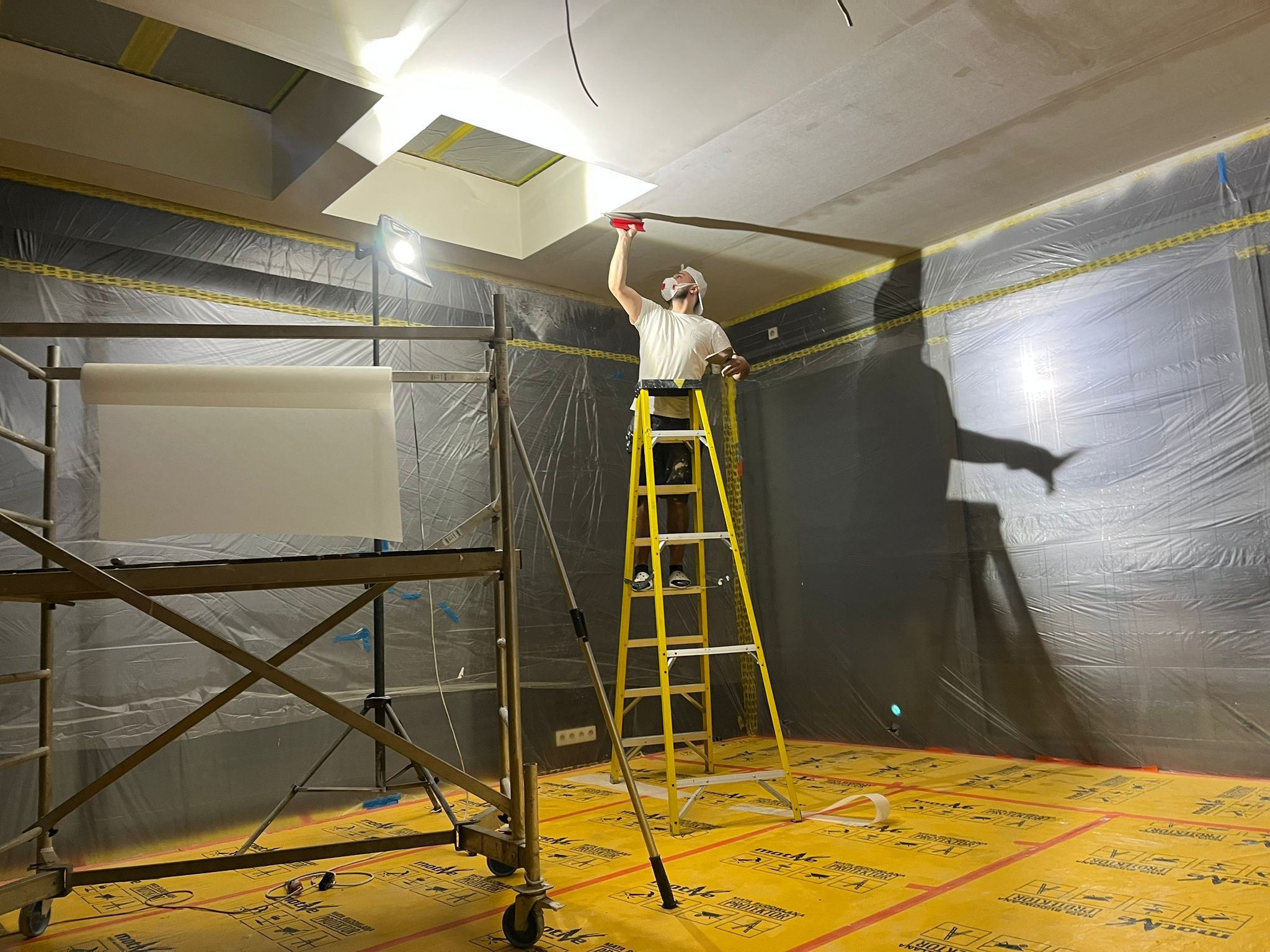 Pracownik na drabinie szpachluje sufit z oknem dachowym. Podłoga i ściany zabezpieczone folią. Widoczne rusztowanie i lampa oświetleniowa.