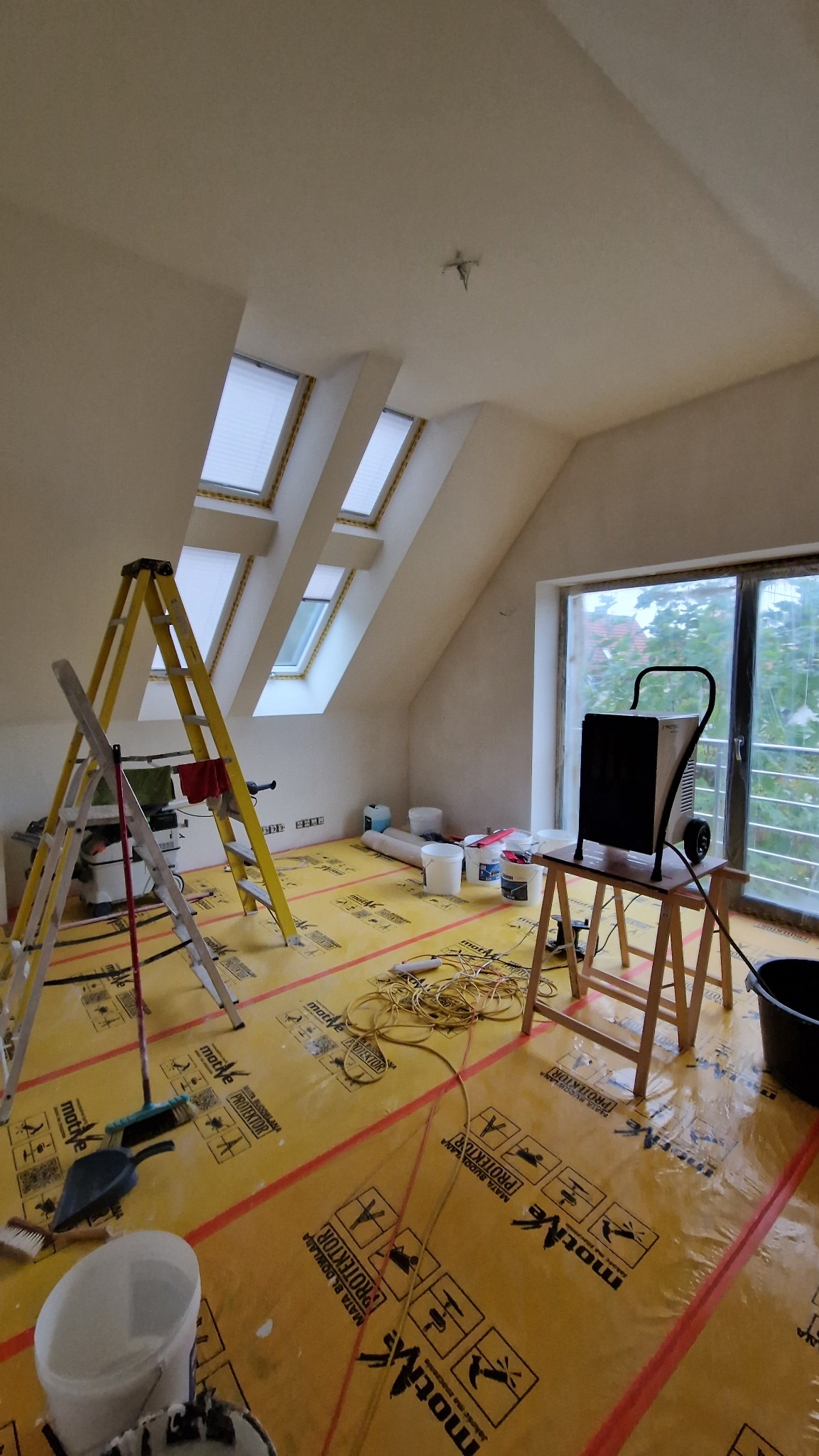 Pokój na poddaszu w trakcie prac wykończeniowych: żółta folia ochronna na podłodze, drabiny, narzędzia i osuszacz powietrza. Widoczne okna dachowe i duże okno balkonowe.