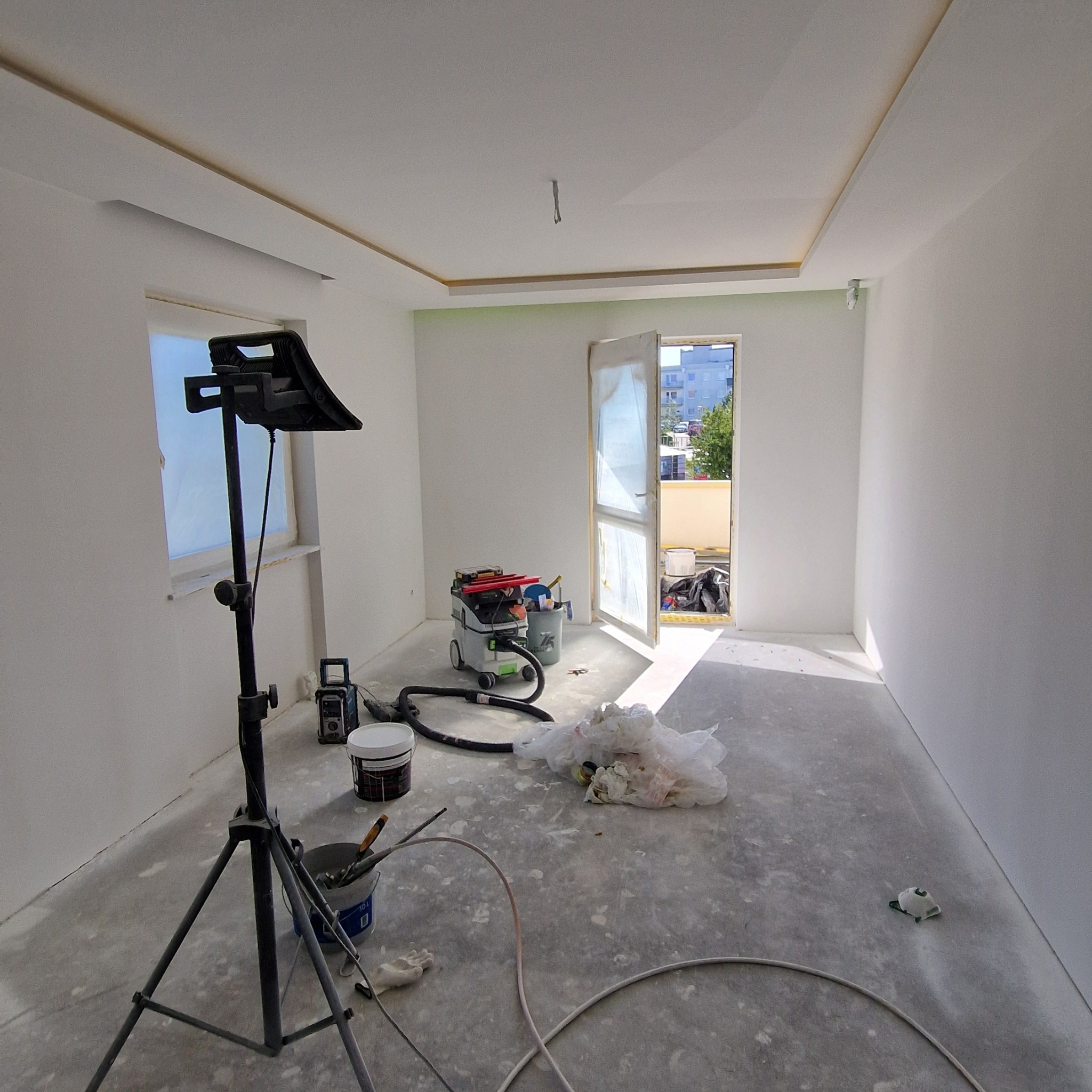 Wnętrze pokoju po gładziowaniu ścian i sufitu, z widocznym oświetleniem roboczym, narzędziami i odkurzaczem przemysłowym. Okno i drzwi balkonowe zabezpieczone folią.