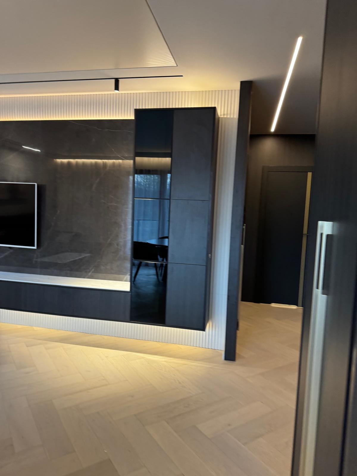 Nowoczesny salon z parkietem w jodełkę, ciemną zabudową ściany z podświetleniem LED i widokiem na kolejne pomieszczenie przez otwarte drzwi.