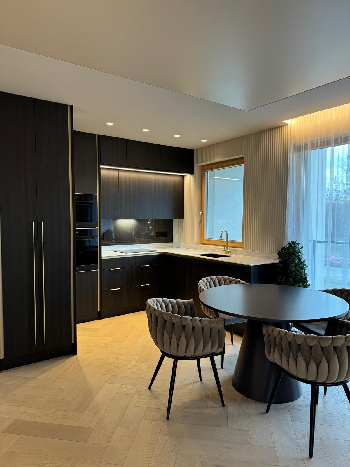 Nowoczesna kuchnia z ciemnymi szafkami, złotymi akcentami i parkietem w jodełkę. Okrągły stół z krzesłami w stylu glamour. Minimalistyczne oświetlenie.