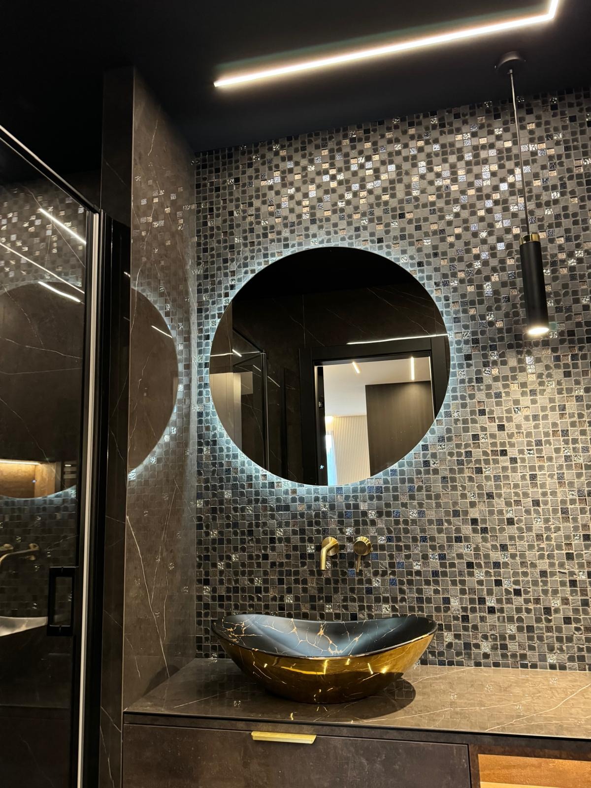 Mozaika w ciemnych tonacjach, okrągłe lustro z podświetleniem LED, złota armatura i umywalka w łazience. Nowoczesny design z akcentami glamour.