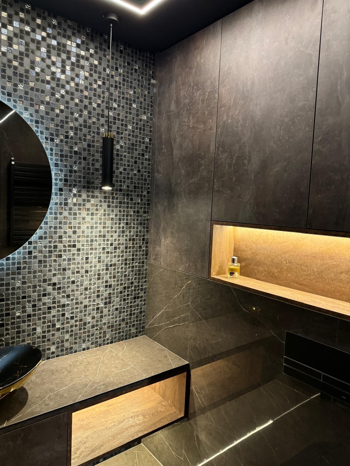 Elegancka łazienka w ciemnych tonacjach: mozaika, marmurowe płytki, podświetlane półki i okrągłe lustro. Nowoczesny design z dbałością o detale.