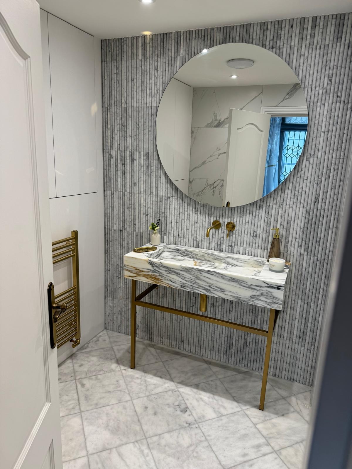 Elegancka łazienka z marmurową umywalką na złotym stelażu, okrągłe lustro i grzejnik drabinkowy w kolorze złota. Płytki marmurowe na podłodze i ścianie.