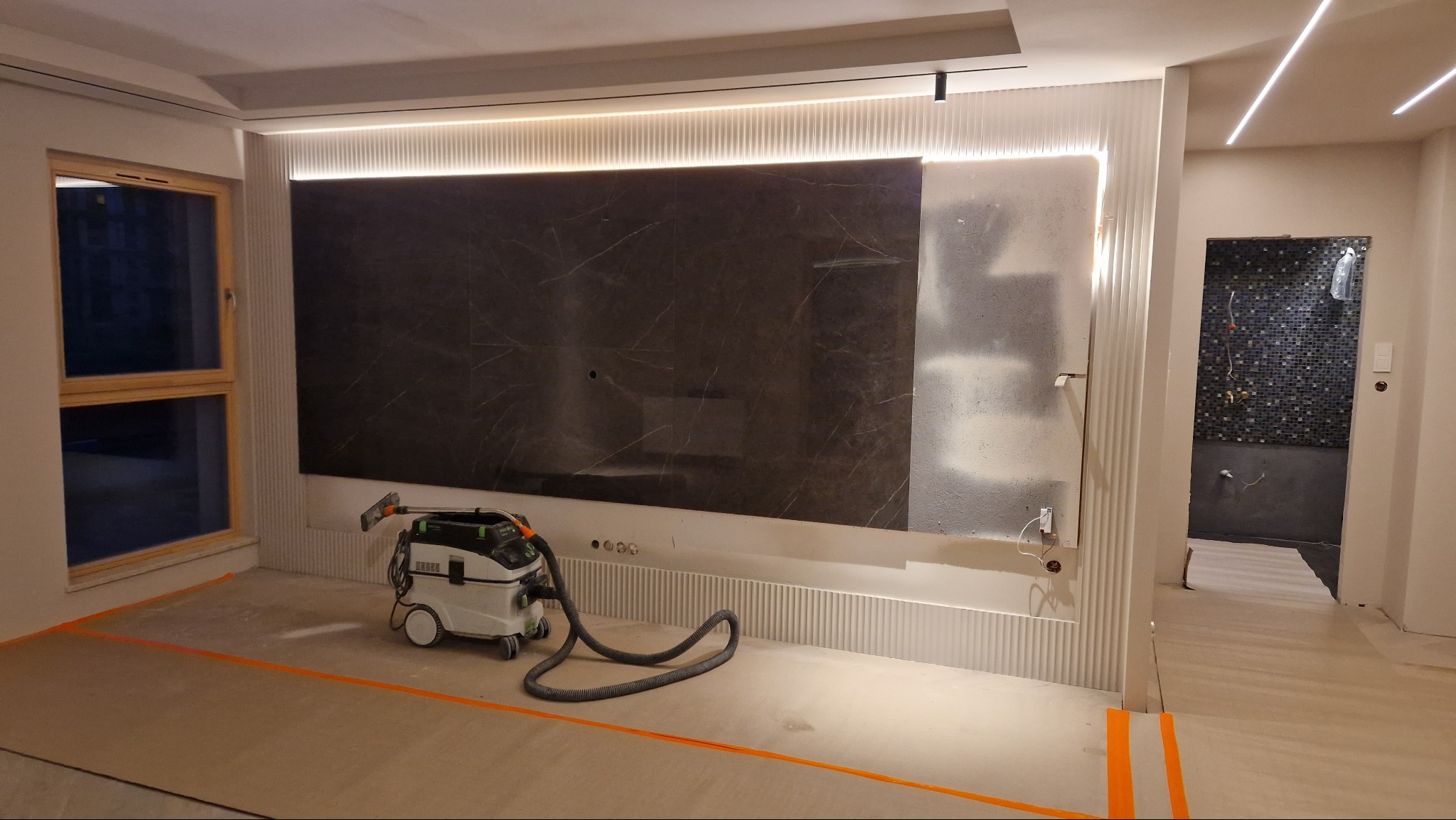 Nowoczesny salon w trakcie prac wykończeniowych: marmurowa ściana TV z podświetleniem LED, widoczny odkurzacz przemysłowy i fragment łazienki z mozaiką.