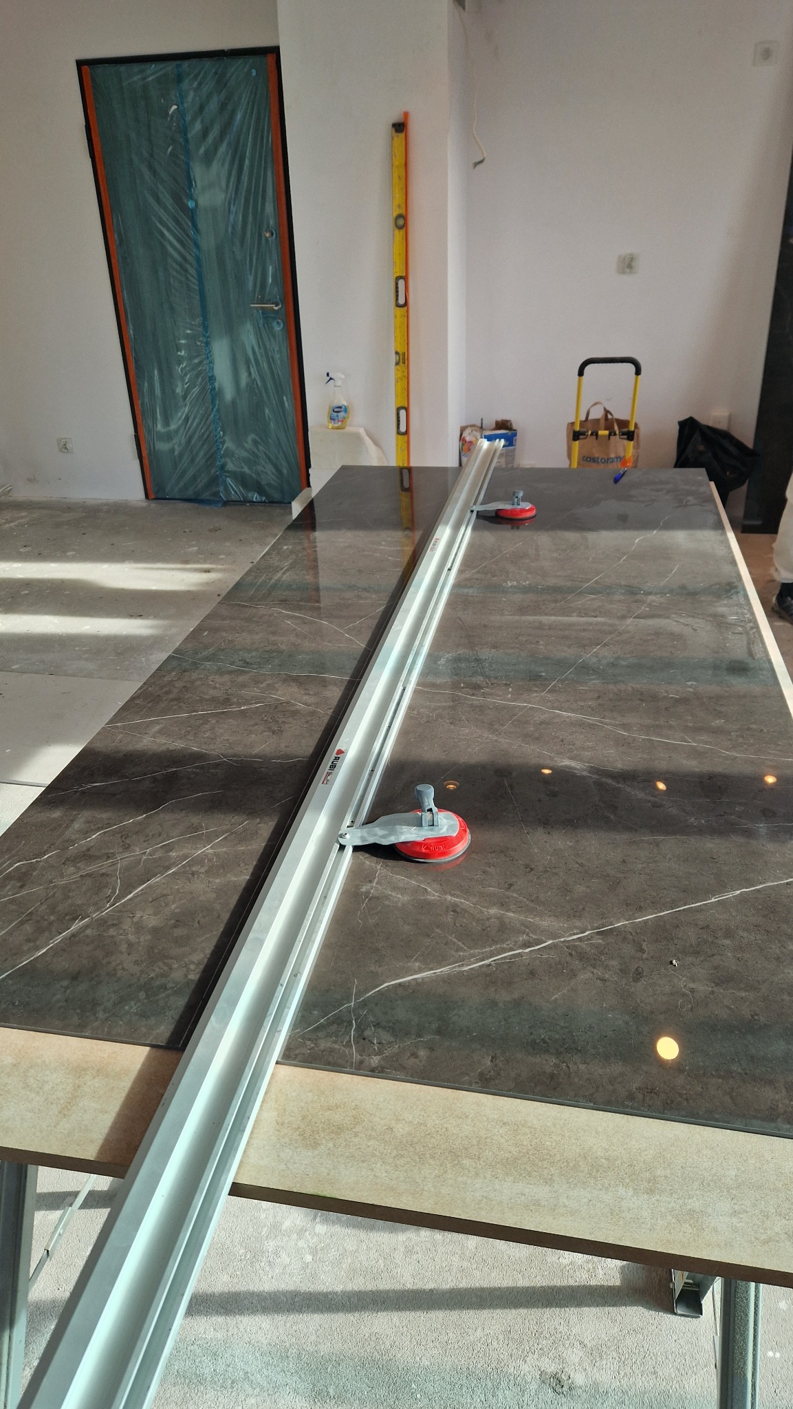 Ciemna, marmurkowa płyta na stole z narzędziem do cięcia płytek Rubi Slim Cutter Plus. W tle poziomica i drzwi zabezpieczone folią.